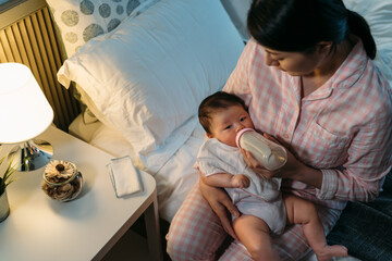 view from top asian mother wearing pajamas is nursing her adorable newborn baby with bottled milk by the bedside table in a cozy bedroom with lamp light.