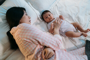 high angle of asian cute tiny baby is lying awake on bed while her mother wearing pajamas is patting her to sleep beside during bedtime in the bedroom at home.