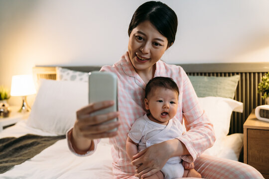 happy asian mother wearing pajamas holding and looking at smartphone screen is taking a selfie of herself and her lovely baby in arms in the bedroom at night.