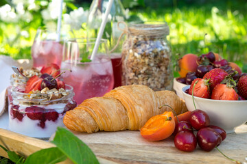 Healthy breakfast of croissant, fruits and berries and parfe with yogurt and granola served on a tray in the garden with green grass and white flowers in the background. 