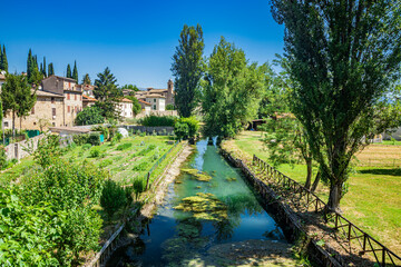 Fototapeta na wymiar The Chiasco river that crosses the ancient medieval village of Bevagna. Perugia, Umbria, Italy. Trees, vegetation, cultivated gardens. Green algae on the surface of the stagnant water.