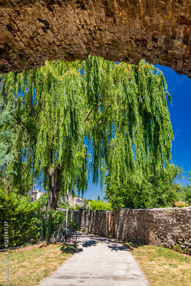 Wall mural A glimpse of the medieval village of Bevagna. Perugia, Umbria, Italy. Blue sky in a sunny summer day. The big weeping willow below the masonry bridge.