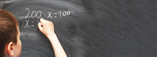 Boy standing back in front of school blackboard and writing. Schoolboy solves math example at the chalkkboard
