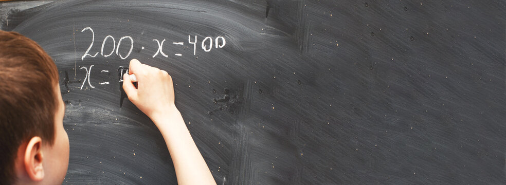 Boy standing back in front of school blackboard and writing. Schoolboy solves math example at the chalkkboard