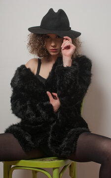 Portrait of a fashionable model with black fur sitting in a green chair in studio. young beautiful woman in black pantyhose .Portrait of attractive woman sitting in chair with black hat