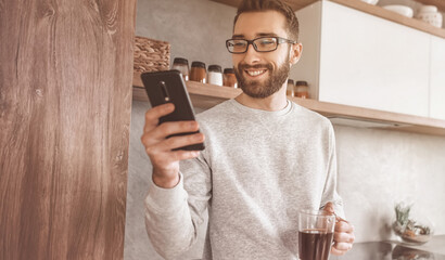 smiling man with smartphone and Cup of coffee standing in his kitchen