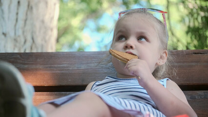 Fototapeta premium Little girl eats gingerbread. Close-up portrait of blonde girl sitting on park bench and eating cookies.