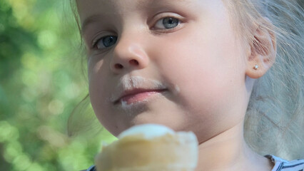 Cute little girl eats ice cream outside. Close-up portrait of blonde girl sitting on park bench and eating icecream.