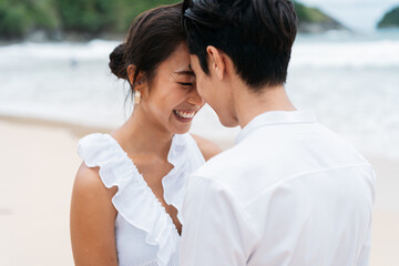 Happy smiling Asian 30s couple in relationship head to head on the beach in summer. A romantic couple wearing a white shirt and dress