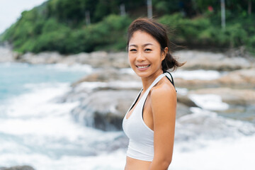 Happy smiling Asian 30s woman looking at camera on the rocky beach in summer with wavy ocean in background. Carefree and calm young lady in summer outfit.