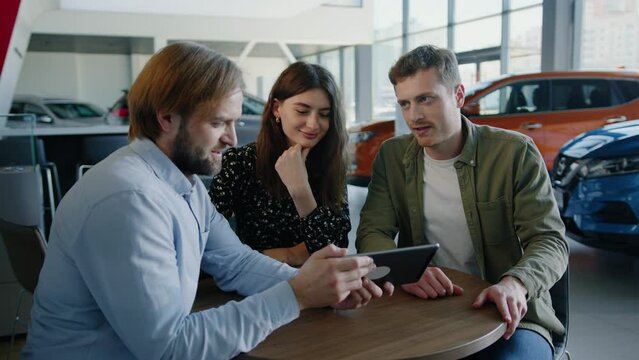 A young couple is looking at options for buying a new car at a dealership on a tablet. The manager sells the car
