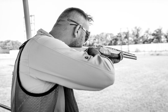 Adult man in sunglasses and a rifle vest practicing fire weapon shooting. Young experienced male aiming shotgun in outdoor.