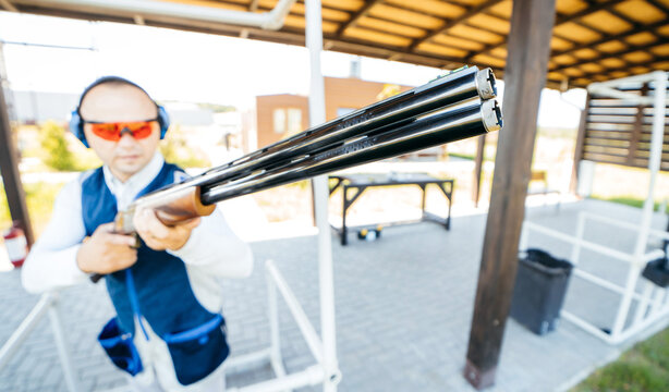 Blurred Background With Focus On Front Of Shotgun Barrel Of Adult Man In Sunglasses, Protective Headphones And A Rifle Vest Practicing Fire Weapon Shooting. Young Experienced Male Aiming At The Camera