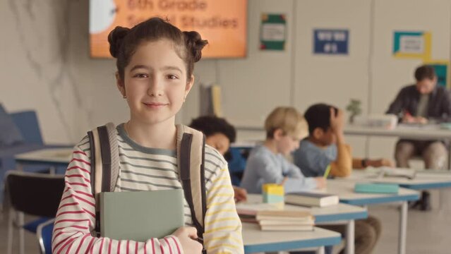 Waist up slowmo portrait of pretty 11 year old Caucasian school girl with backpack and books in hands smiling to camera in classroom