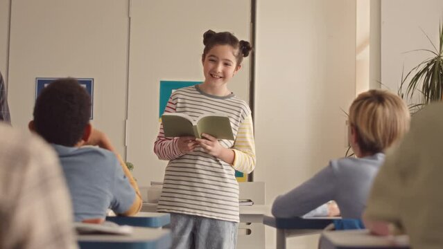 Slowmo pf pretty Caucasian school girl in striped longsleeve holding book while making report in front of teacher and classmates during lesson