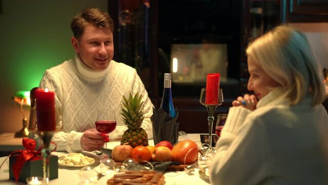 Portrait of loving man toasting sitting at table dining with shy woman. Happy smiling Caucasian adult boyfriend dating with girlfriend at home indoors. Valentine's celebration concept