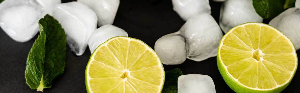 high angle view of melting ice cubes near sliced limes and milt leaf on black background, banner.