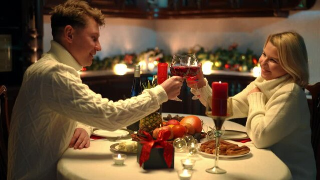 Side view portrait of man and woman toasting clinking glasses drinking red wine on Valentine's Day. Medium shot of happy loving Cauacsian couple celebrating holiday at home indoors talking