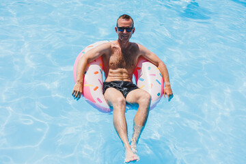 A young man in sunglasses and shorts is relaxing on an inflatable donut in the pool. Summer vacation - obrazy, fototapety, plakaty