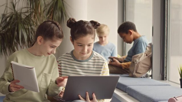 Medium slowmo of two girls using laptop while making school project or homework in school corridor. Boys chatting on windowsill in background