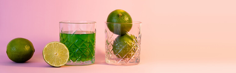 green alcohol drink near faceted glass with fresh limes on pink background, banner.