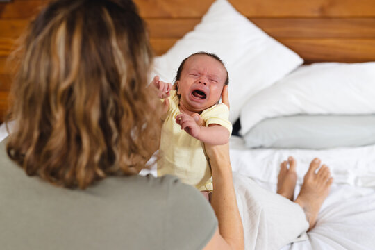 Mid adult caucasian mother consoling crying newborn baby while sitting on bed at home, copy space