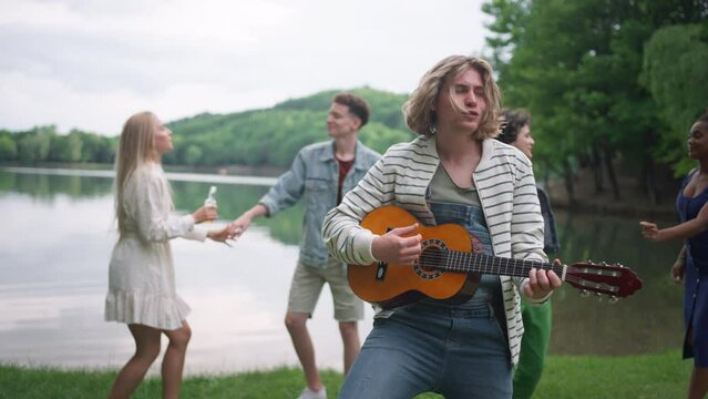 Group of young friends having fun on picnic near a lake, dancing, singing and playing guitar.