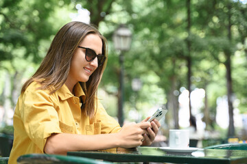 Side view at smiling young woman using smartphone outdoors, texting online during coffee break in summer cafe, positive smiling lady in sunglasses and yellow casual shirt spends time in social network