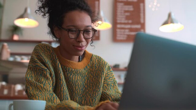 Positive woman wearing yellow sweater and eyeglasses working on laptop in cafe