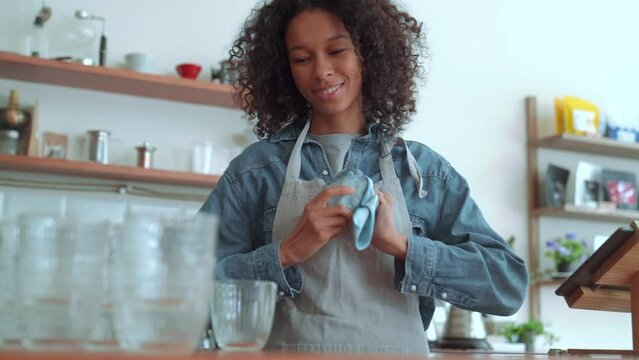 Positive woman barista wiping glass cups while standing in cafe