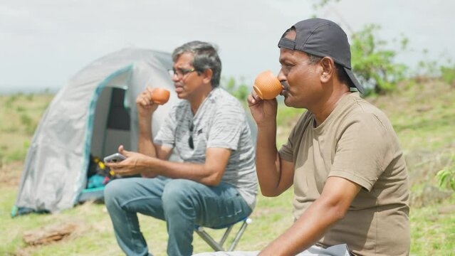 Middle aged hikers having tea while talking each other near camping tent at destination during weekend holidays - concept of refreshment, friendship and travellers.