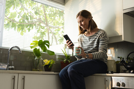 Low angle view of caucasian mid adult lesbian woman holding coffee and using smartphone in kitchen