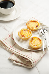 Traditional portuguese vanilla pudding puff pastry pastel de nata on black oven rack on white plate on checkered kitchen towel with a cup of coffee in white porcelain