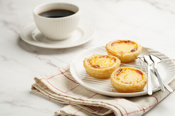 Traditional portuguese vanilla pudding puff pastry pastel de nata on black oven rack on white plate on checkered kitchen towel with a cup of coffee in white porcelain