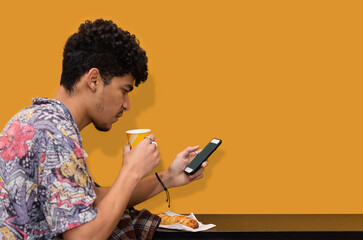 Latin man with curly hair and wearing a colorful shirt having a hot drink while checking his cell phone isolated on yellow background
