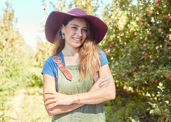 A beautiful female farm worker standing with her arms crossed on a fruit farm during harvest season. Young happy farmer between fruit trees in summer. Agricultural industry growing produce