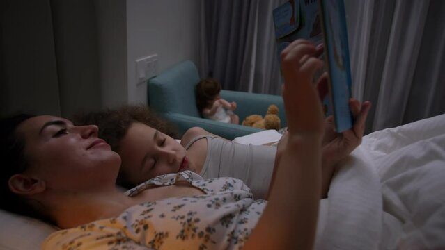 Child care at home, Mom takes care of the youngest daughter, reads a bedtime story to the children, Free time for children. Mother reading book to daughter while lying in bed at home