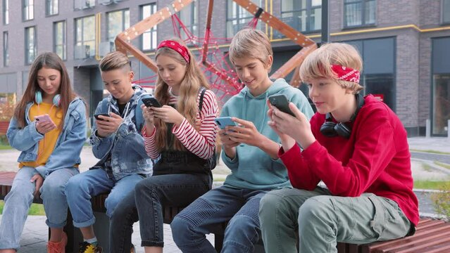 Smiling group of teens using gadgets sitting in row on bench. Happy boys and girls users obsessed with modern technology devices holding smartphones, tech addiction, teenagers concept