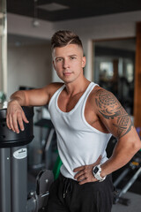Handsome muscular man athlete model with hairstyle in a white tank top mock up stands and poses near the simulator at the gym