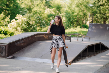 Teenager girl with a skateboard and holding cup of coffee walking on the skatepark