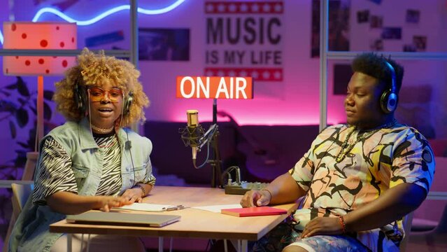 African American male and female podcast hosts in headphones smiling and talking on camera while sitting at table in recording studio with colorful neon light