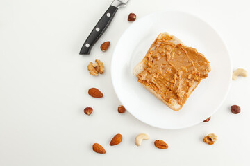 Bread with peanut butter on a plate. Knife and various types of nuts. Cashew, almond, walnut, hazelnut isolated on white background.