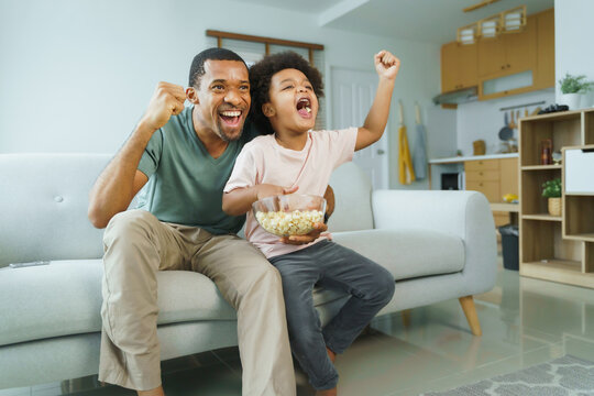 Cheerful African American Father and his little boy watching and cheering sports games competition together at home, celebrate