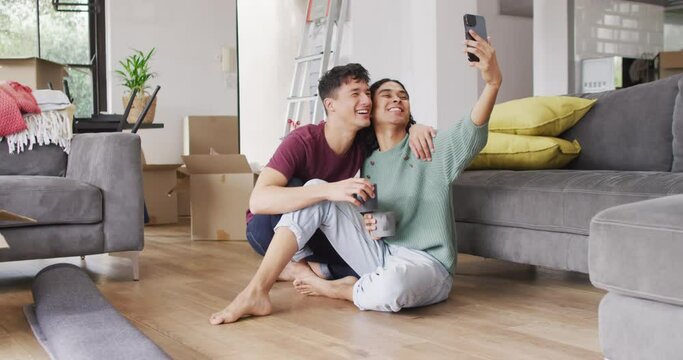 Happy diverse male couple moving house, drinking coffee, taking selfie using smartphone