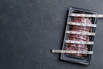 Pack of raw sliced Dry Aged beef in black plastic tray and waist tape on black stone table background. Food and healthcare concept - obrazy, fototapety, plakaty