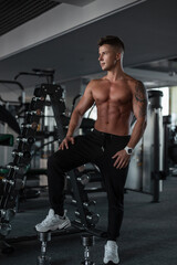 Obraz na płótnie Canvas Handsome young athletic guy with hair and a bare muscular torso stands and poses near the dumbbells in the gym