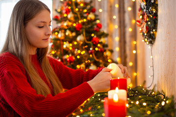 Young blond girl in red sweater igniting candles in decorated living room. Preparations for Christmas