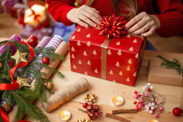 Close-up shot of a big box present wrapped in festive wrapping paper on table, girl sticking big bow on top of it. Christmas presents preparations