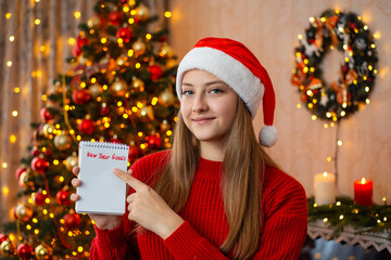 Young beautiful girl in red Santa hat with notes pointing finger to inscription New Year Goals. Concept of wish list or to do list for the comming year