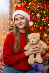 Fototapeta na wymiar Girl with teddy bear near decorated Christmas tree. Portrait of an attractive girl in Santa hat with teddy bear